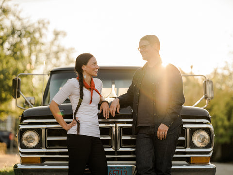 Amanda and Erik stand facing each other and smiling leaning against a vintage truck. The sun shines behind them. 