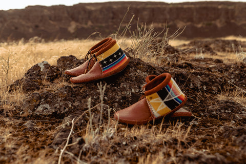 Two pair of moccasins made of leather and brightly pattered wool fabric sit in a grassy field