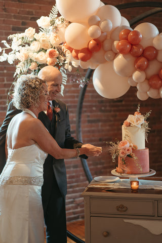 a bride and a groom slicing a piece of cake after their wedding ceremony featuring balloons in peach and burnt orange and a floral piece attached to the balloon garland