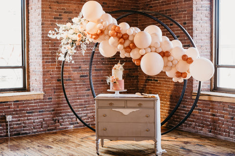 cake on top of small antique-style table, large balloon garland, clustered balloons with floral spray on a circular arch.  Wedding setup for a cake cutting - balloons are peach and burnt orange. Space features large windows and open brick walls.