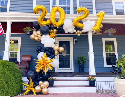black, white and gold balloons clustered to make a balloon garland on a front porch of a house.  Balloons feature gold mylar starbursts balloons and large 2021 balloons in gold