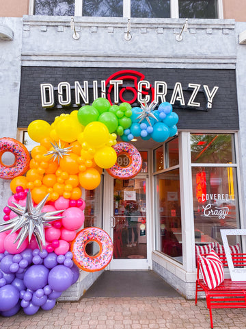 purple, pink, goldenrod, yellow, lime green, and blue balloons clustered to make a balloon garland in front of a retail donut shop in Connecticut.  Balloon garland has large pink frosted donut balloons attached with sprinkles, along with silver starburst balloons