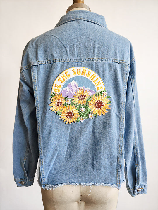 Image of the back of a light blue denim jacket on a dress form in front of a white background.  The denim jacket has a large embroidered patch sewn onto the back of the jacket.  The patch says BE THE SUNSHINE in yellow letters arched over a nature scene of purple snow capped mountains and a field of large yellow sunflowers.