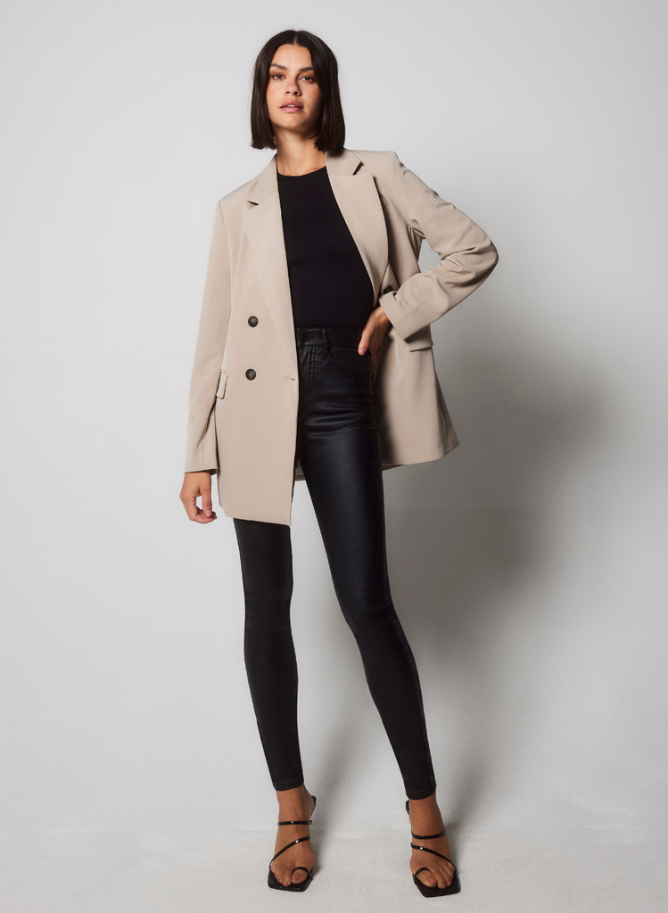 Model wears the Coated Jeans with a black leotard and a beige blazer. She stands against a white background in strappy heels.