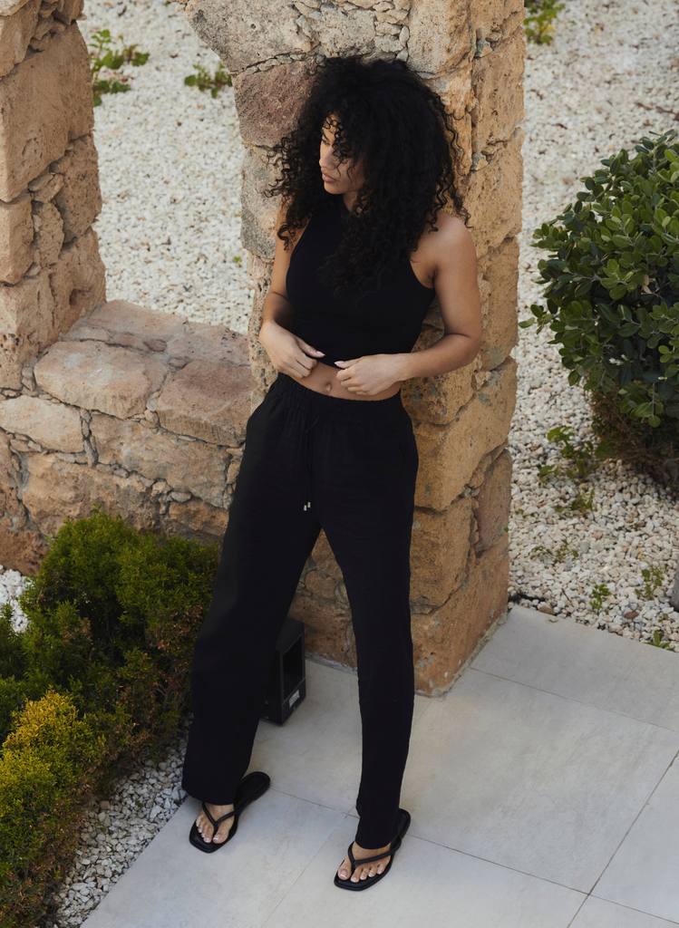 Cyprus setting, woman wears the Linen Black Trousers whilst leaning against a wall in the sunny Cyprus sun.