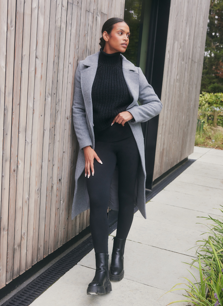 Woman stands outside, wearing the Winter Everyday Leggings in Black. She has styled the outfit with a smart black jumper and smart grey coat.