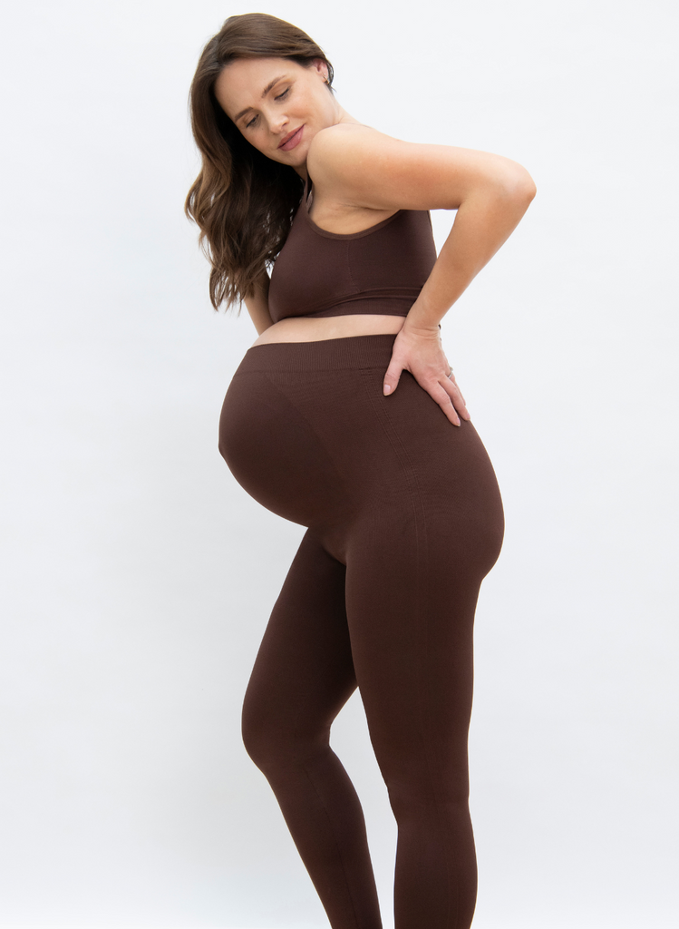 Pregnant woman stands again white backdrop, wearing the Maternity Seamless Set in Chocolate Brown.
