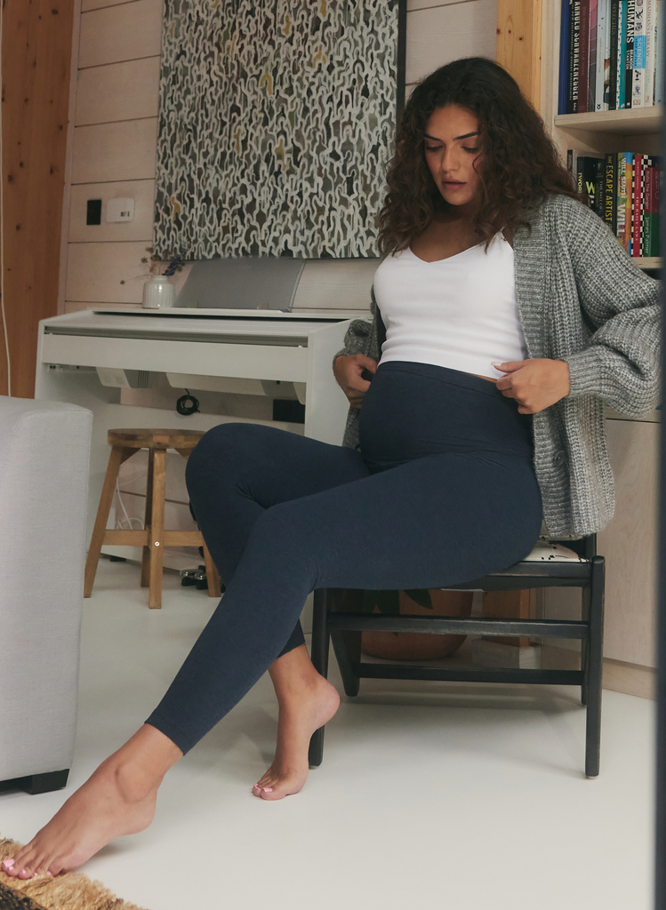 Pregnant woman sits on a chair wearing a white top, cardigan and the Steel Blue Everyday Maternity Leggings.