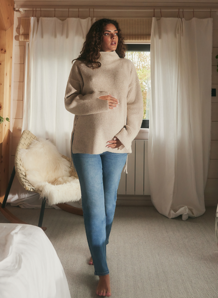 Pregnant woman wears the Maternity Straight Jeans in Light Blue and a cosy jump. She holds her bump, standing in a lounge area.