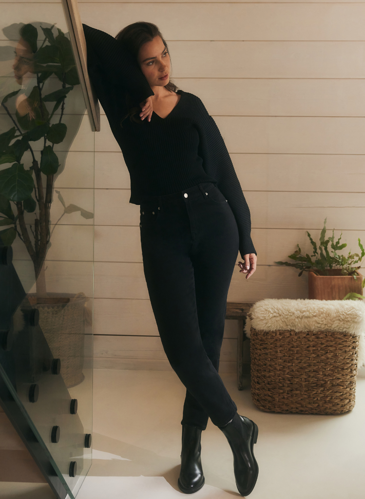 Woman leans against staircase wearing Black Straight Jeans, boots and a black cardigan.