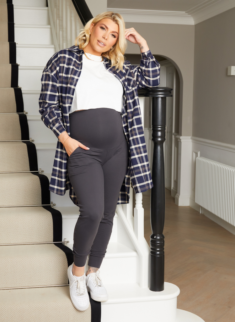 Olivia Bowen wears Maternity Recharge Joggers, with white tops and oversized shirts. She stands on the stairs in her home.