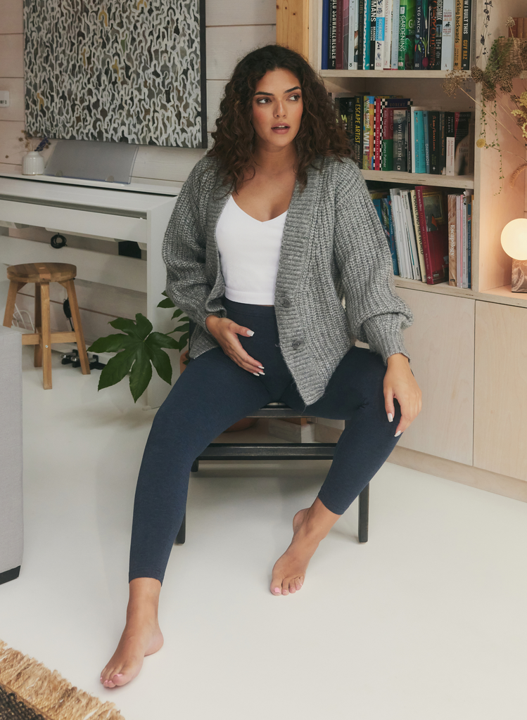 Woman sits in living area, wearing Blue Maternity Everyday Leggings, a white top and a grey cardigan. She holds her baby bump.