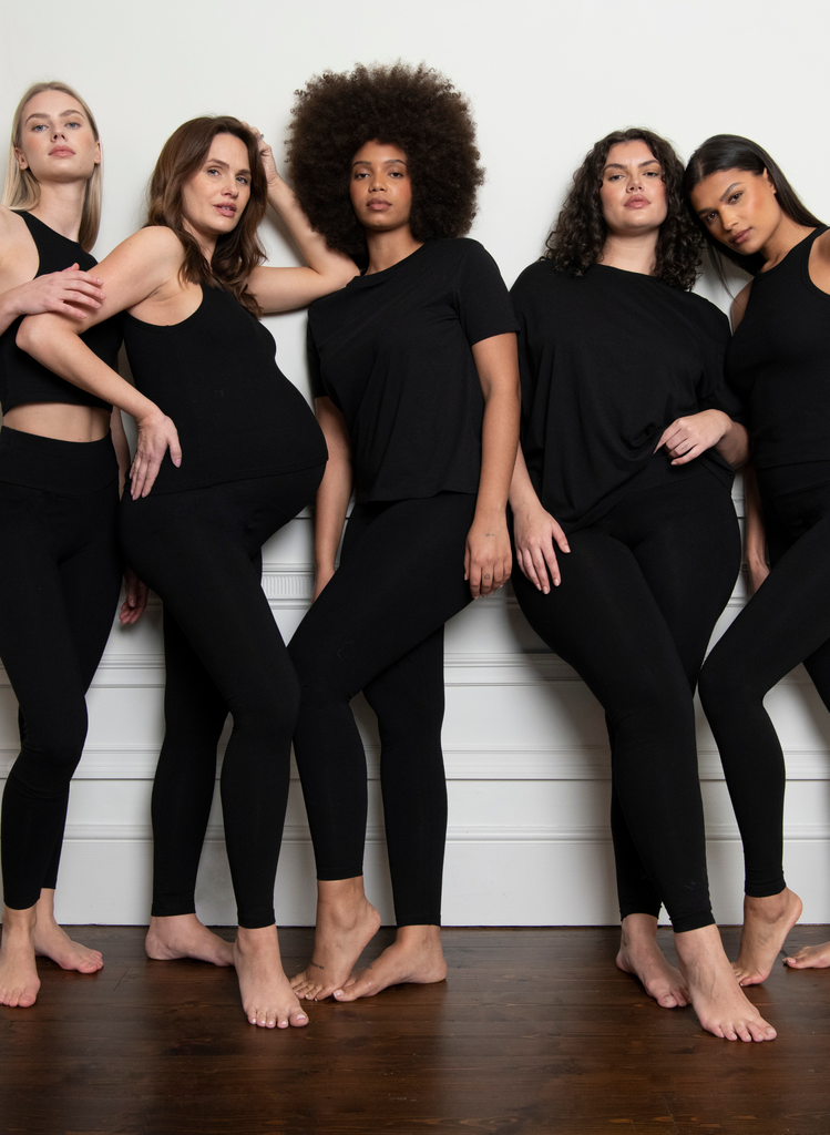 5 Women wear all black and stand against a white wall, posing for the camera. One women is also pregnant.