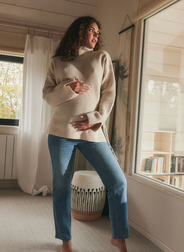 Woman stands in a lounge area, where Straight Maternity Jeans and a knitted jumper. She holds her pregnant stomach and looks out a window.