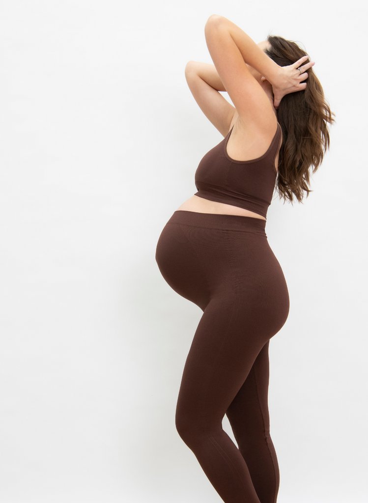 Model wears the Seamless Maternity Leggings in Chocolate Brown, which the matching Bralette. She stands against a white background and faces side on.
