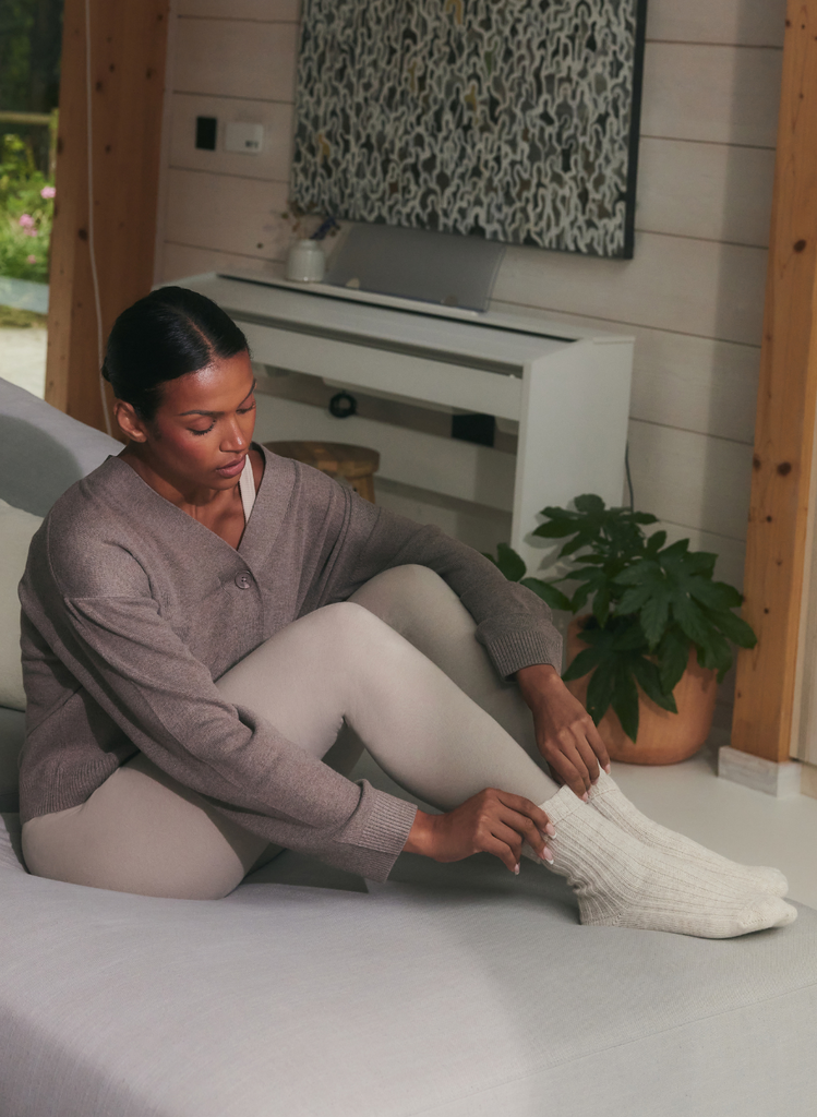 Model sitting on floor in LOVALL Everyday Leggings in Beige, a cosy cardigan, and pulling up some thick, white socks.