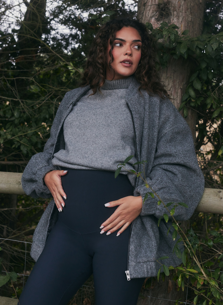Pregnant woman wears in Maternity Winter Leggings in Navy, with a grey jumper and grey jacket. She stands outside, holder her bump.