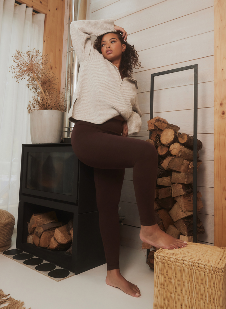 Curve model wears the Chocolate Brown Everyday Leggings and stands next to a log burning fire and logs.