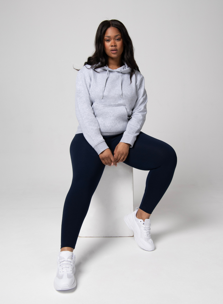 Woman sits on white box, against a white backdrop, wearing the Everyday Curve Leggings in Black and the Everyday Hoodie in Grey.
