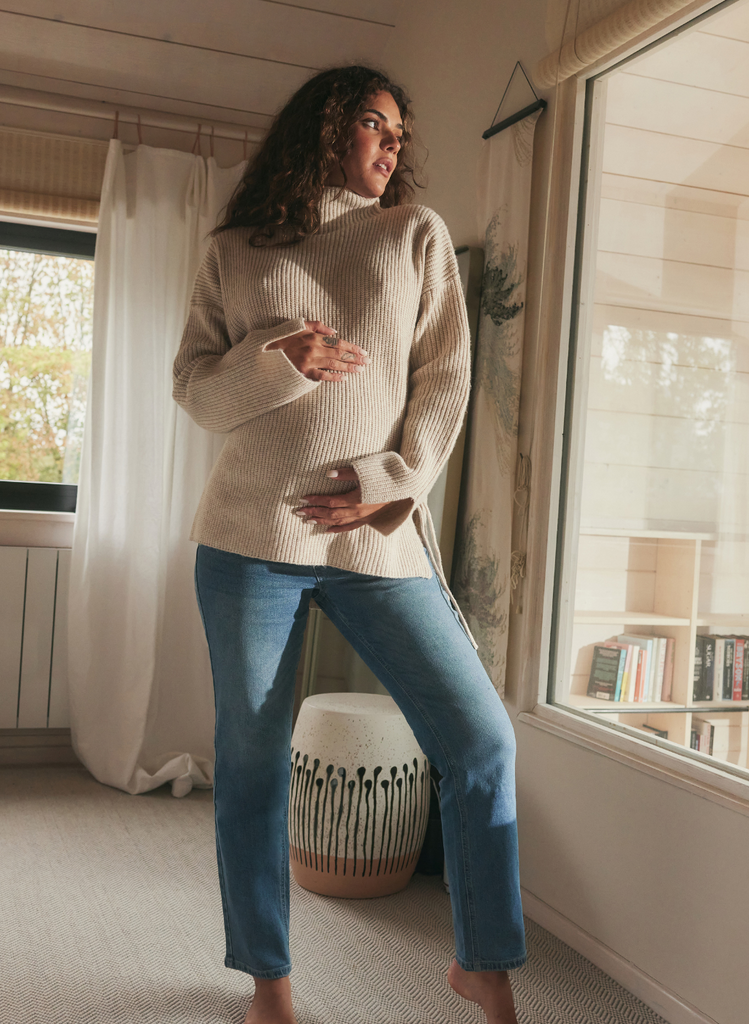 Woman poses with her babay bump in a bedroom setting, next to a large window. She is wearing a chunky knit jumper and Maternity Straight Jeans by LOVALL.