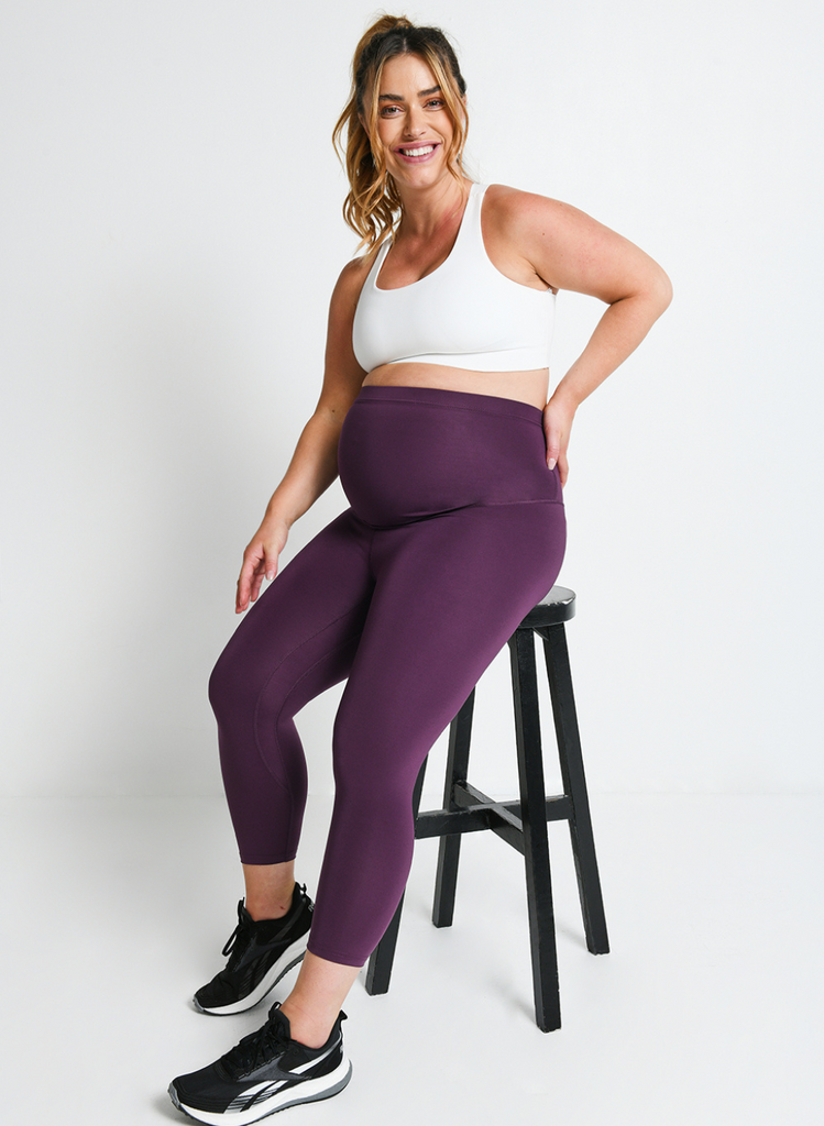 Pregnant model wears the Maternity Revitalise Leggings whilst sat on a stool against a white backdrop.