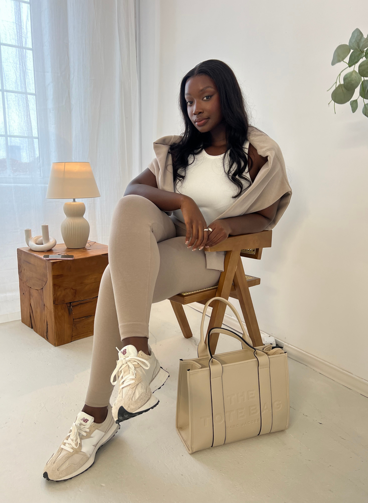 Woman wears Oatmeal Beige Everyday Leggings, the White Ribbed Tank Top, and the Beige Sweatshirt around her shoulders. She sits on a chair next to a Beige Bag and side table.
