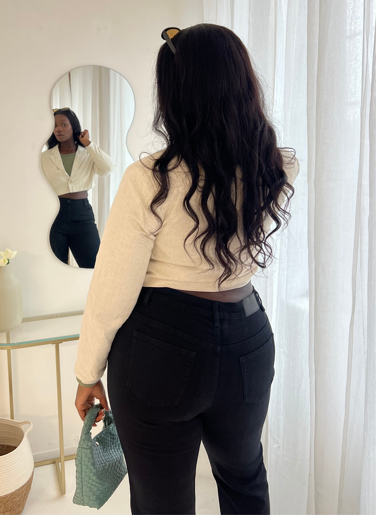 Model wears Black Lift & Shape Jeans, with sage green crop top, and beige cropped blazer. She stands looking in the mirror.