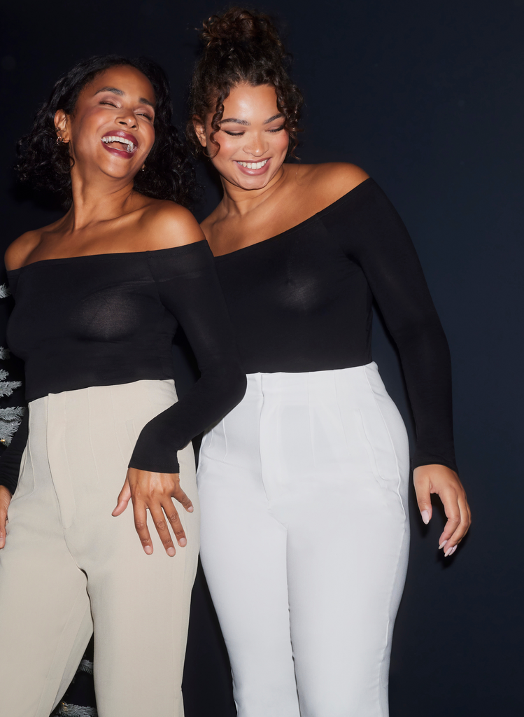Two Models wear the Beige and White Cigarette Trousers, with an off-the-shoulder black top. They stand together laughing.