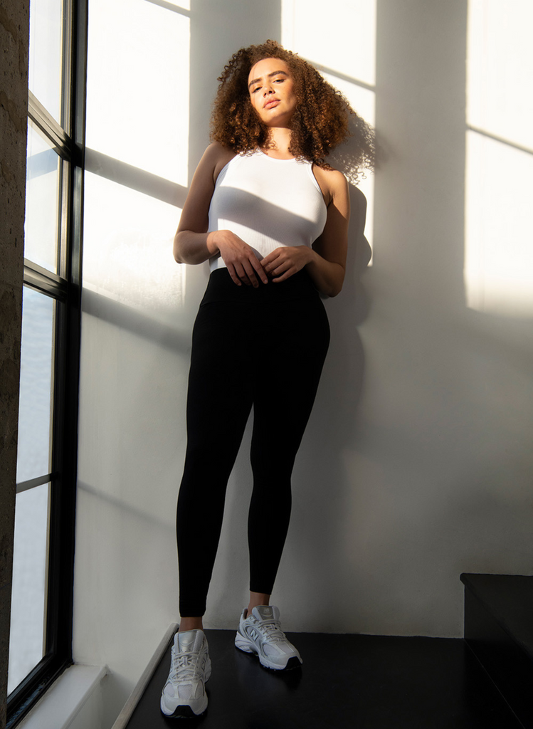 Woman leans against a wall in the light of a window, wearing the White Ribbed Tank Top and Everyday High Waisted Leggings in Black.