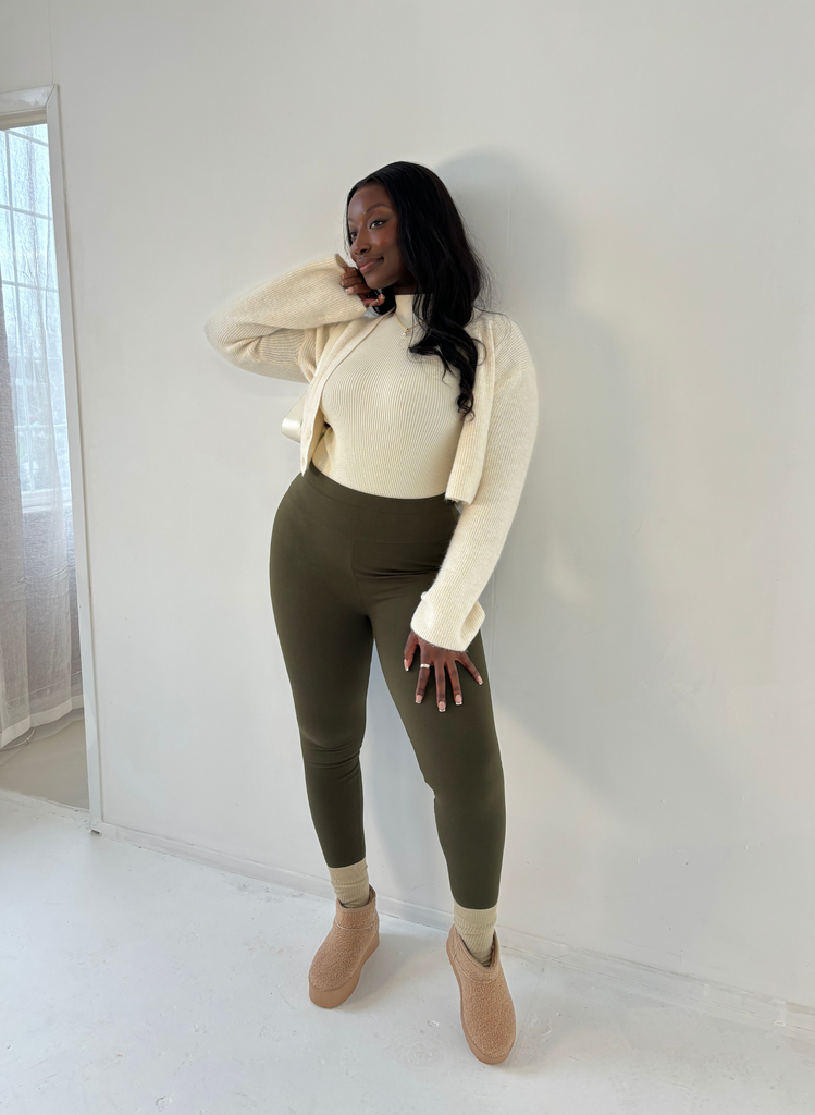 Woman poses against a white wall wearing the Khaki Green Everyday Leggings, with a white turtle neck and cardigan. She also wears Ugg shoes and fluffy socks.