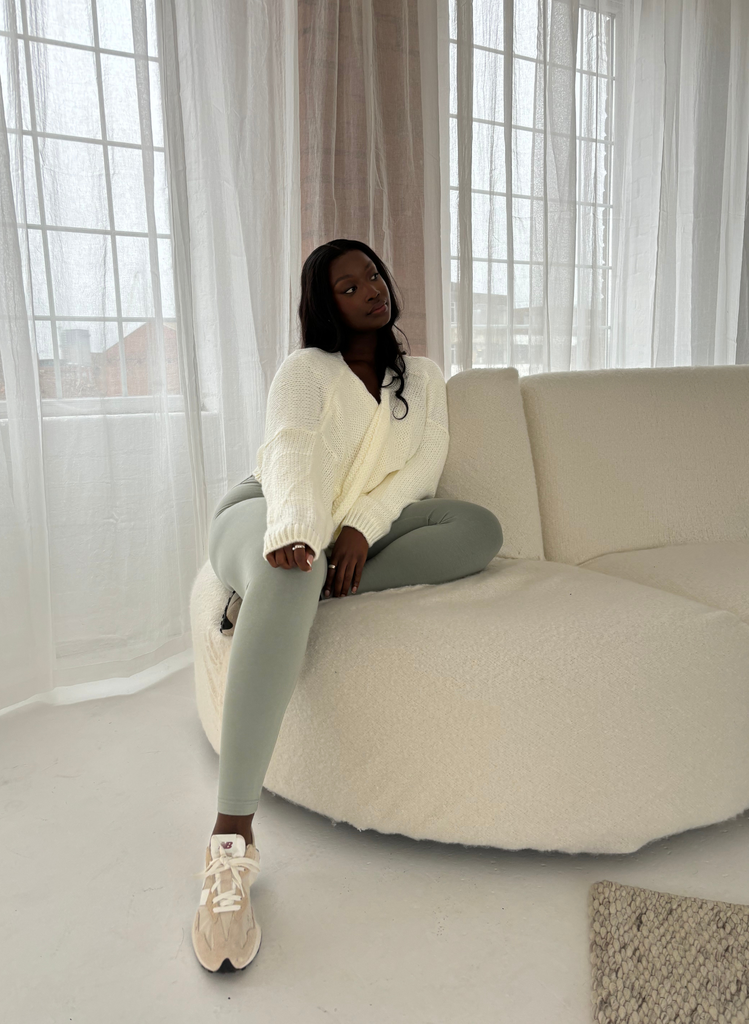 Woman wears the Sage Green Everyday Leggings with a white cardigan and trainers. She sits on a white sofa in a lounge area.
