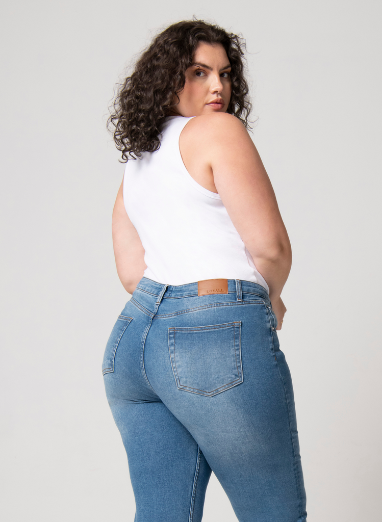 Woman wears white tank top with mid blue mom jeans. She faces the back to the camera with her head over her shoulder.