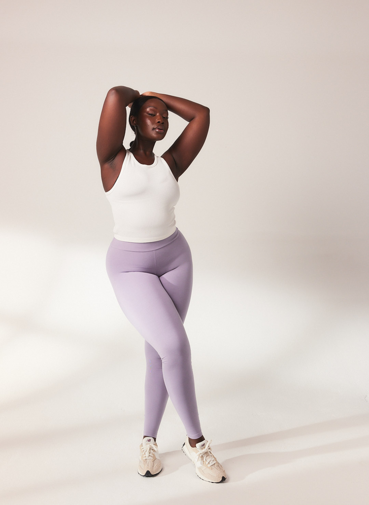 Woman stands in a studio, her arms are above her head and her eyes are closed. She wears Mauve Lightweight Leggings, with a white crop top and white trainers.