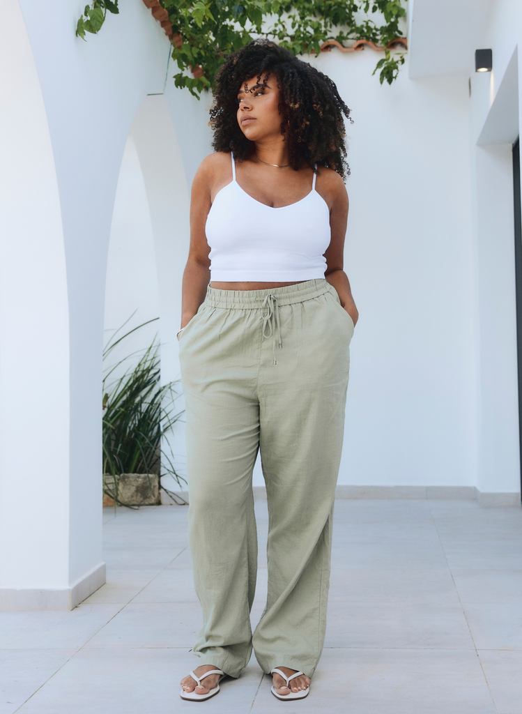 Model wears Sage Green Linen Trousers with white crop top. She stands outside with her hands in her pockets.