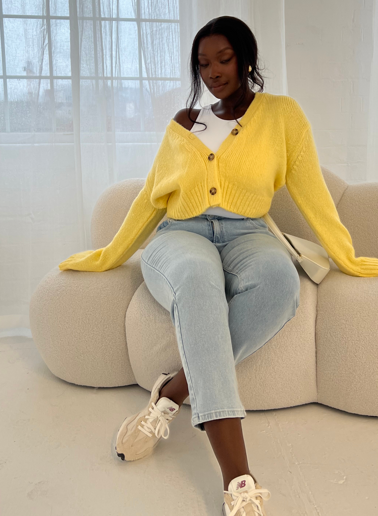 Model wears light bleach Jeans with yellow cardigan and sits on cream sofa.