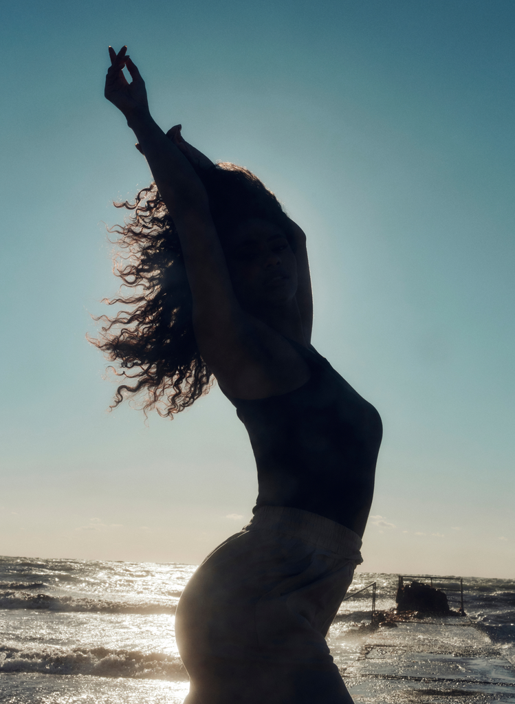 Woman stands on the beach and the photo captures her silhouette, as she stands with her arms in the air and hair blowing in the wind.