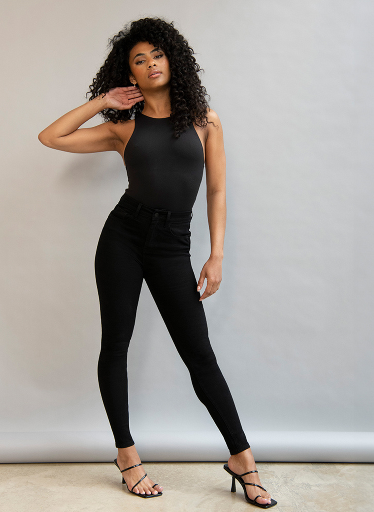 Woman poses wearing black skinny jeans from the Lift & Shape collection. She's paired them with a black leotard and heels.