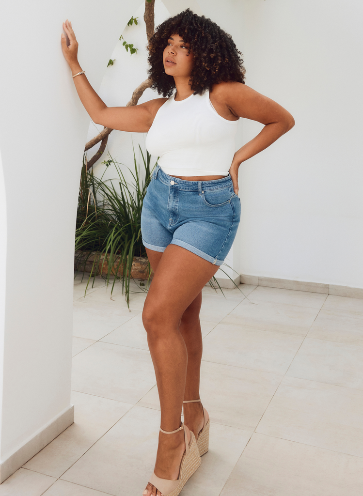 Woman poses in Denim Shorts and a white top, wearing heeled sandals in the outdoor sunshine of Cyprus. 