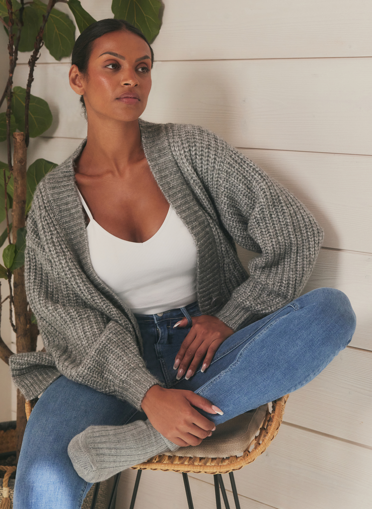 Woman sits on a chair wearing the mid blue skinny jeans from the Lift & Shape collection. She's paired them with a grey cardigan, grey socks and a white tee.
