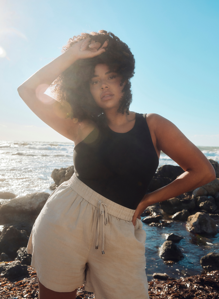 Woman wears Stone Linen Shorts with Black Tank Top, standing in front of the sea.