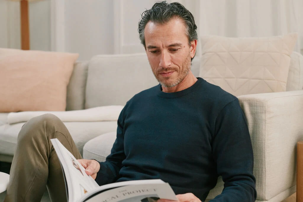 Model reads a newspaper in a living room wearing the Asuwere Cotton Silk Crew knitted sweater in a Navy Colour