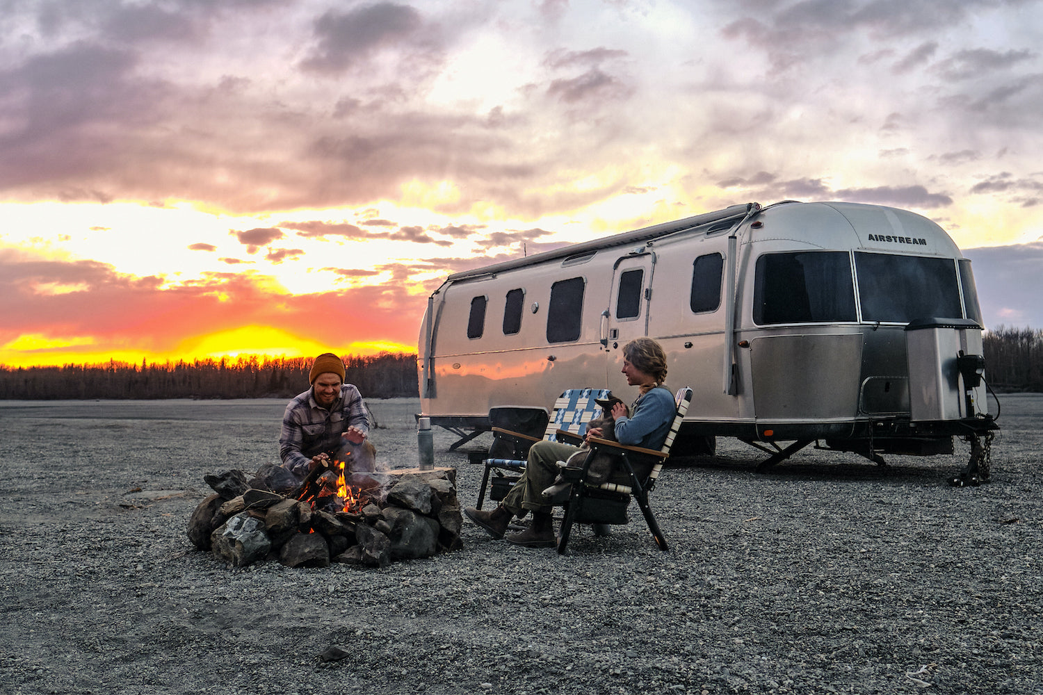 Collin & Kendal Strachan sitting in voyager outdoor chairs in Alaska with their airstream and a sunset in the background.