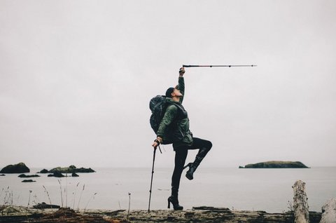 With platform heels and hiking poles, Pattie poses on a driftwood log in front of a vast grey body of water