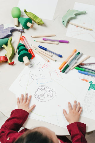 child drawing on a table with art products and wooden toys