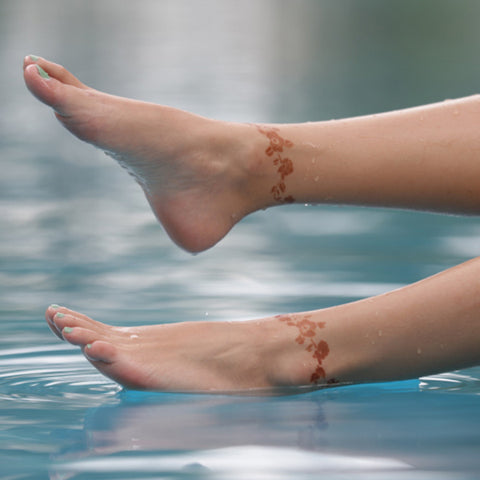 A woman enjoys the water, submerging her feet, adorned with an azalea henna tattoo.