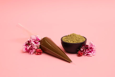 An image showcasing a bowl of henna powder along with a henna cone & decorated with flowers.