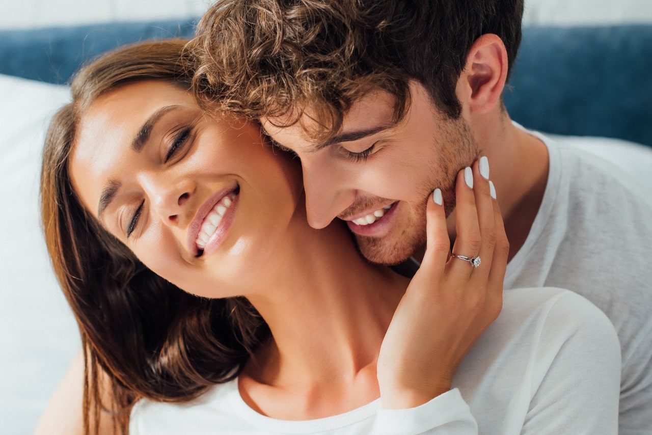 A man and woman cuddling together with the woman’s hand touching the man’s face and showing off her engagement ring.