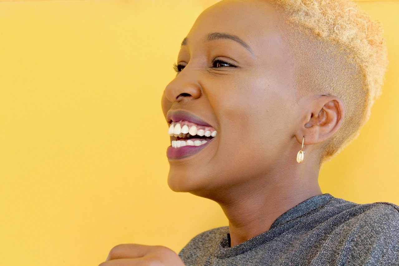 A woman with blonde hair and purple lipstick smiles against a yellow background.