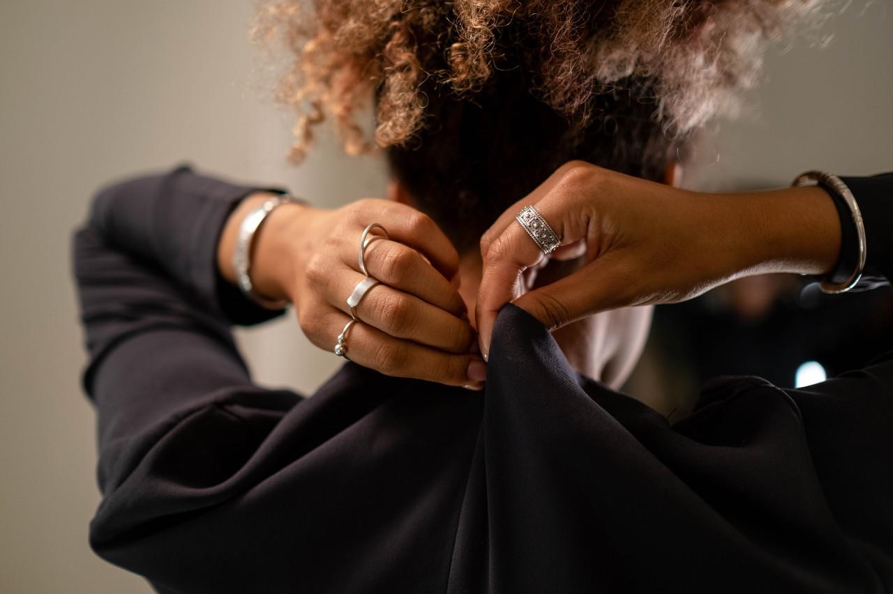 A woman buttoning the top button on the back of her black shirt, showing off multiple sterling silver rings and a bangle on each arm.