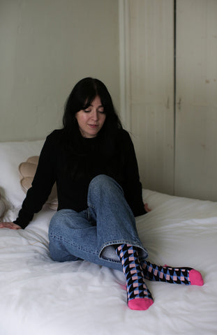 Besma from Curiously Conscious sitting on a bed wearing black Dimensional recycled cotton socks, blue jeans and a black cardigan